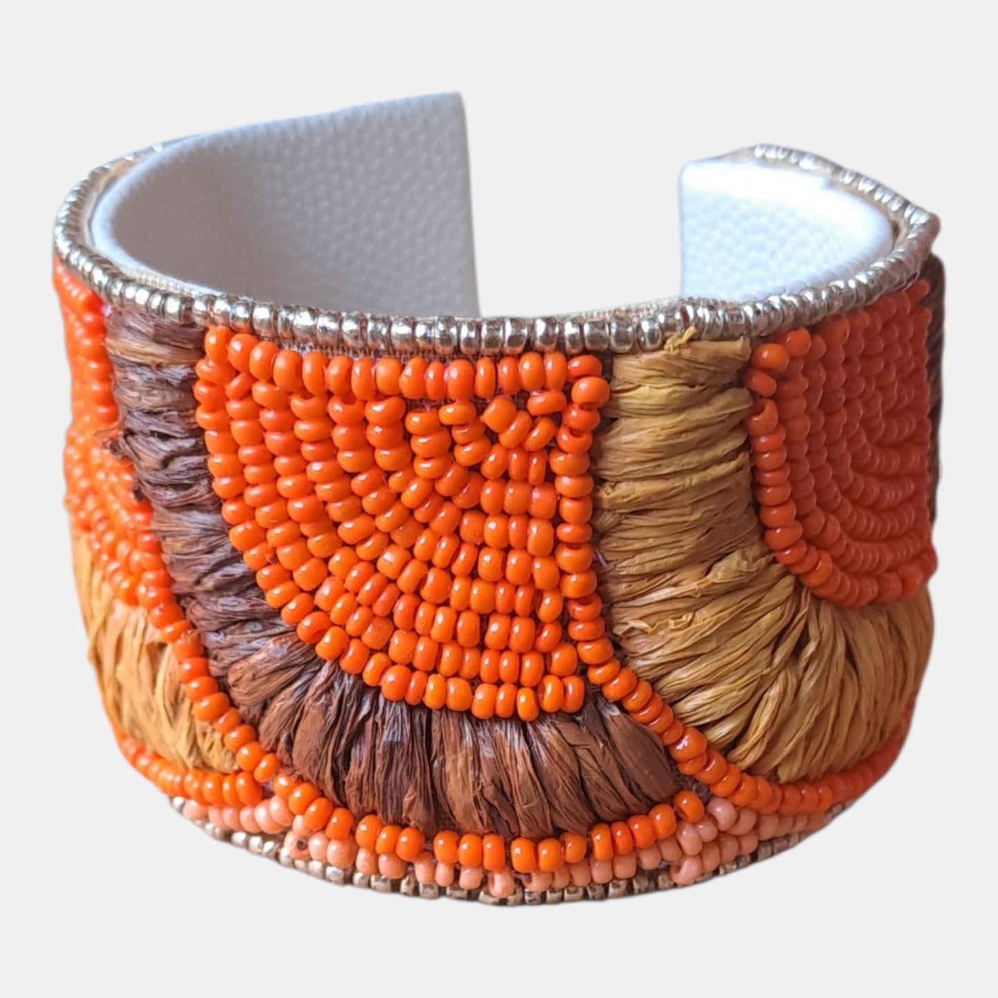 A wide cuff bracelet featuring geometric patterns of bright orange, brown, and tan seed beads over a white textured inner lining, finished with silver-toned beaded trim, measurements: 4,5cm wide, 18cm circumference