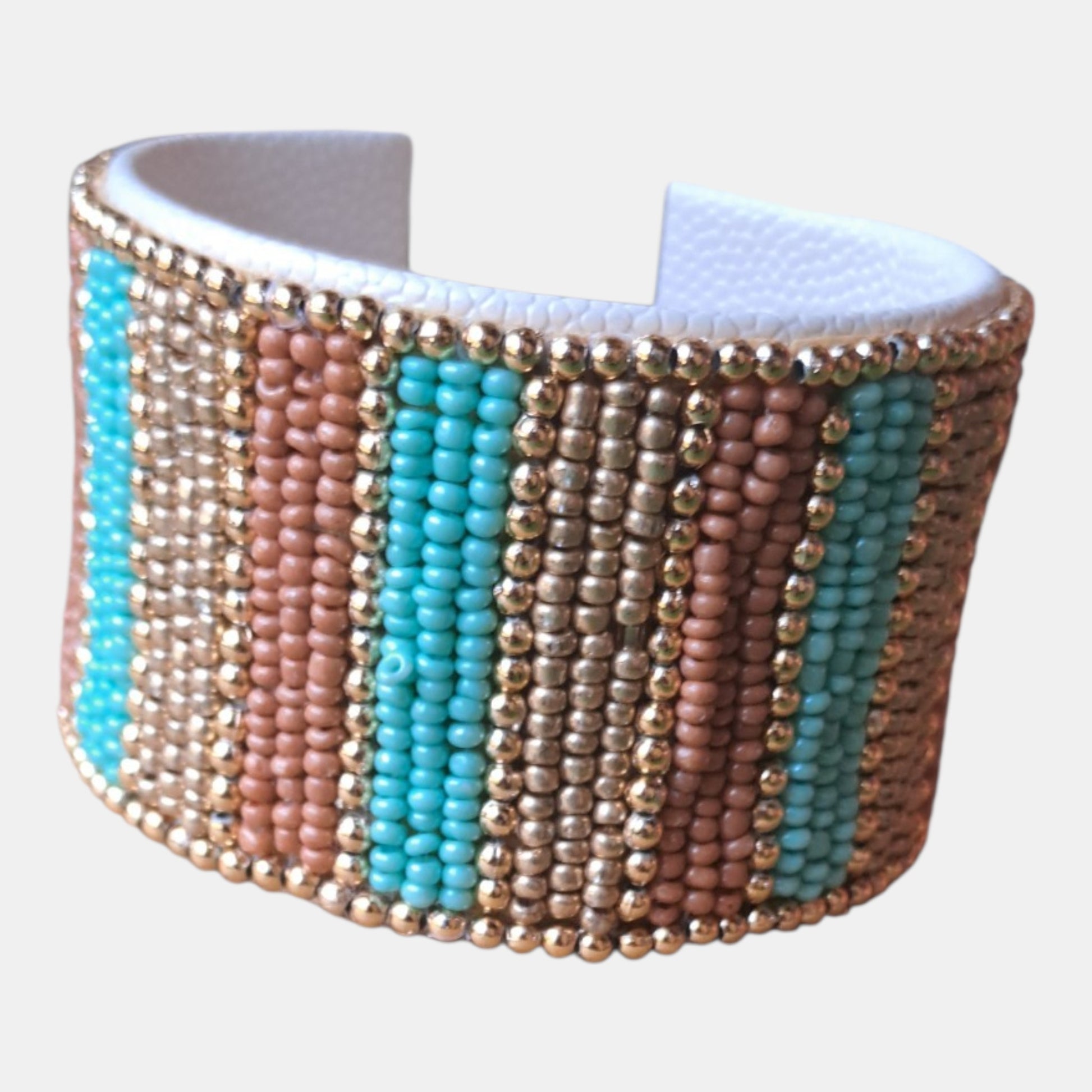 A wide cuff bracelet featuring vertical stripes of turquoise, dusty pink, and metallic gold seed beads, with a white faux-leather interior, Measurements: 4,5cm wide, 18cm circumference