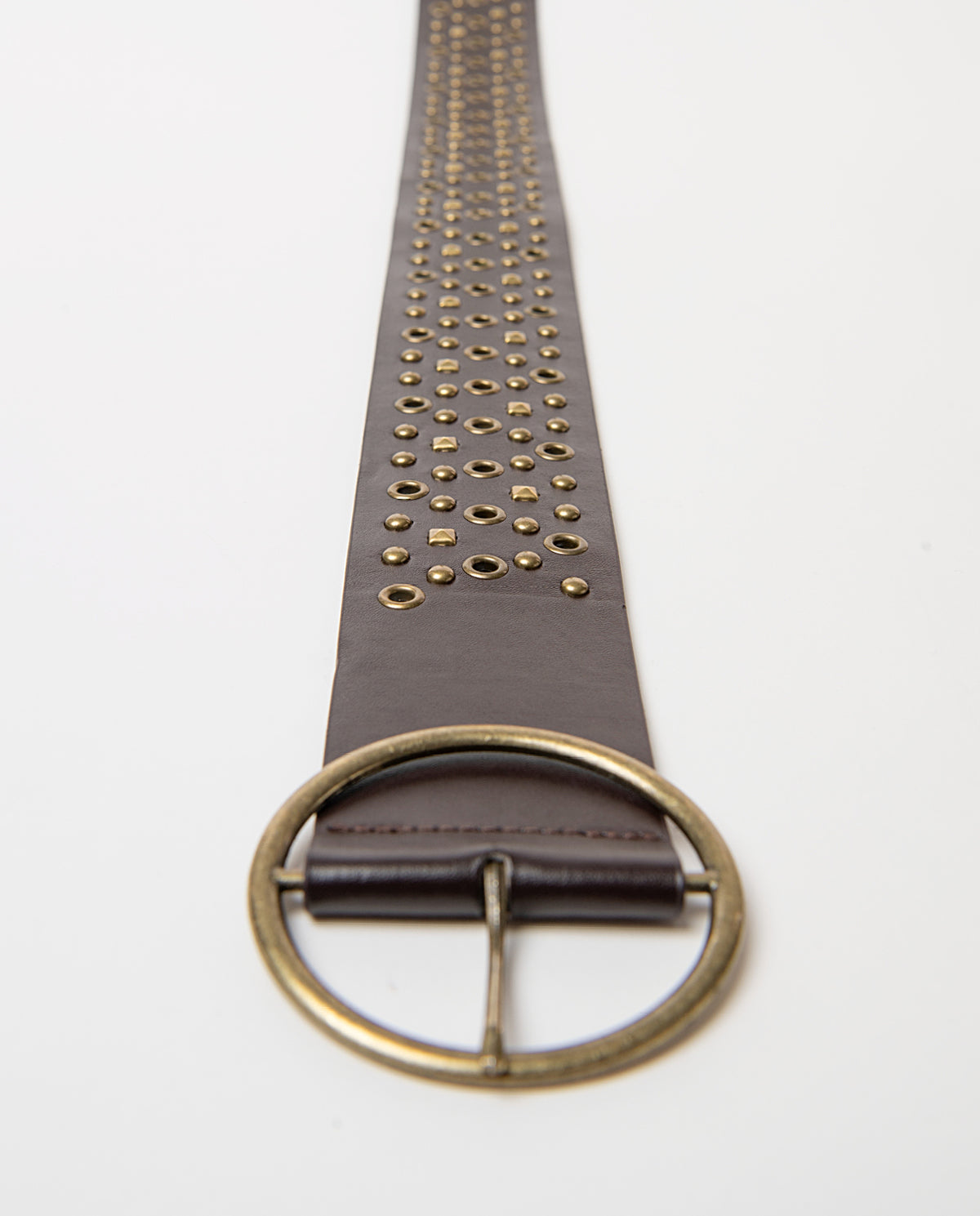 Wide brown belt in smooth faux leather, covered in studs and metal eyelets with an aged finish, and a round buckle. 104cm long, 6.5cm wide