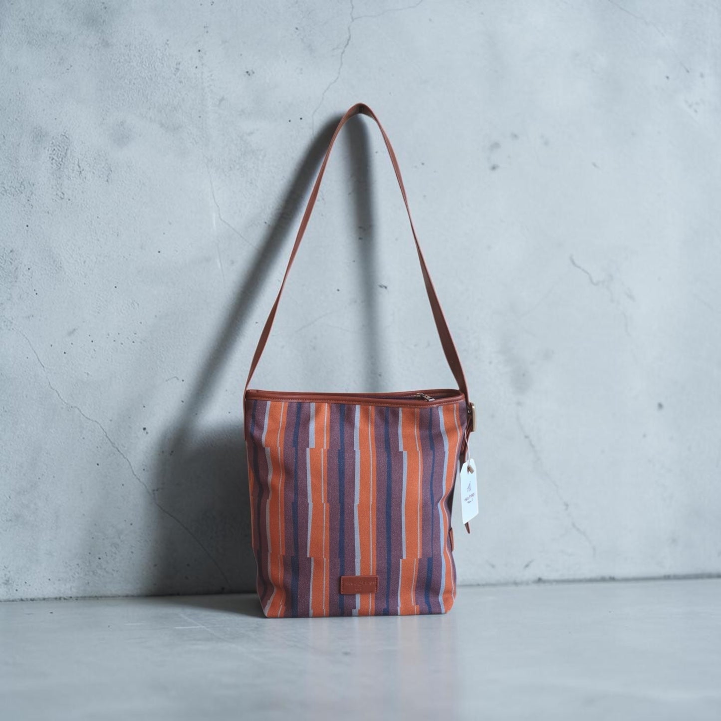 A striped canvas shoulder bag with vertical orange, purple, and blue stripes, brown leather trim and strap, composition: 95% Cotton,5% Polyurethane,measurements: 33 cm tall x 26 cm wide x 14 cm deep.