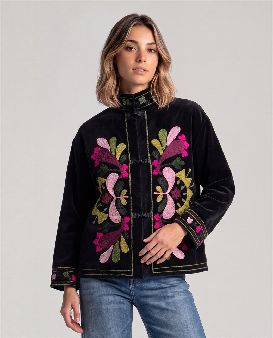 Short black velvet jacket with a mandarin collar and knot fastenings. Colorful, symmetrical flower embroidery gathers at the center front, with detailing at cuffs and hem. Long sleeves.
