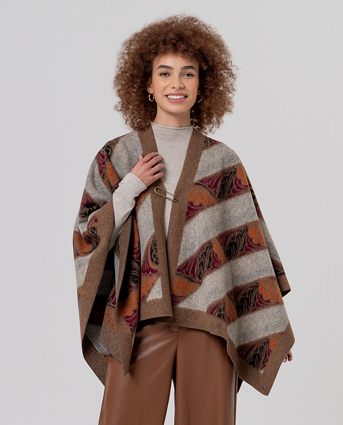 Open beige cape-poncho in warm knit with a pin fastening and contrasting trim. Wave and abstract jacquard in brown, grey mustard and bordeaux colors with a felted texture.