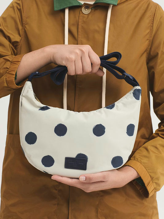 Printed polyester ecru curved crossbody bag with navy dots print, zip closure and adjustable navy strap, inside pocket, debossed logo, 8 cm tall x 26 cm wide x 7 cm deep.