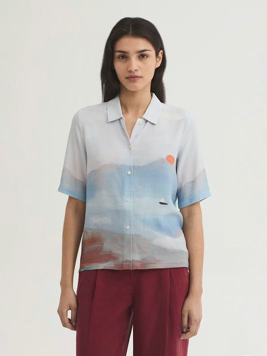 Penelope panneau print shirt in light blue, turquoise, dusty blue color background viscose crêpe, with  a watercolor design of a small boat into the sea in a red sunset, shirt collar, button front and short sleeves.