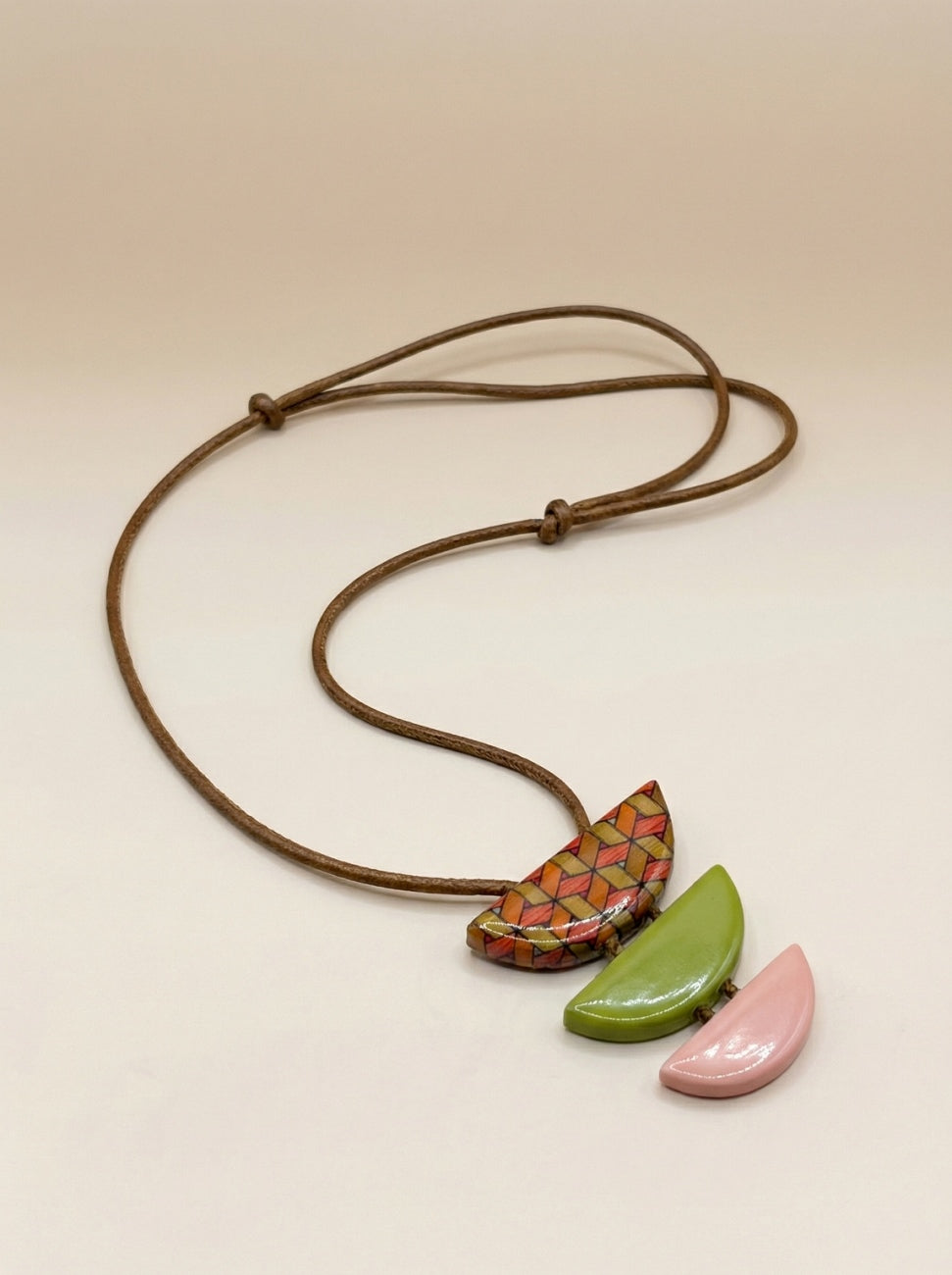 A handmade-style necklace with a brown adjustable cord and three distinct pendant shapes: a central multi-colored geometric patterned pendant, a glossy green leaf-shaped pendant, and a matte pink leaf-shaped pendant