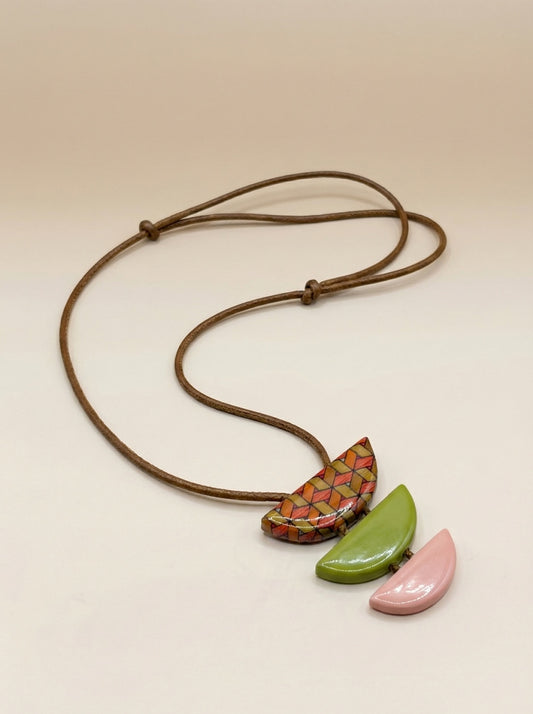 A handmade-style necklace with a brown adjustable cord and three distinct pendant shapes: a central multi-colored geometric patterned pendant, a glossy green leaf-shaped pendant, and a matte pink leaf-shaped pendant