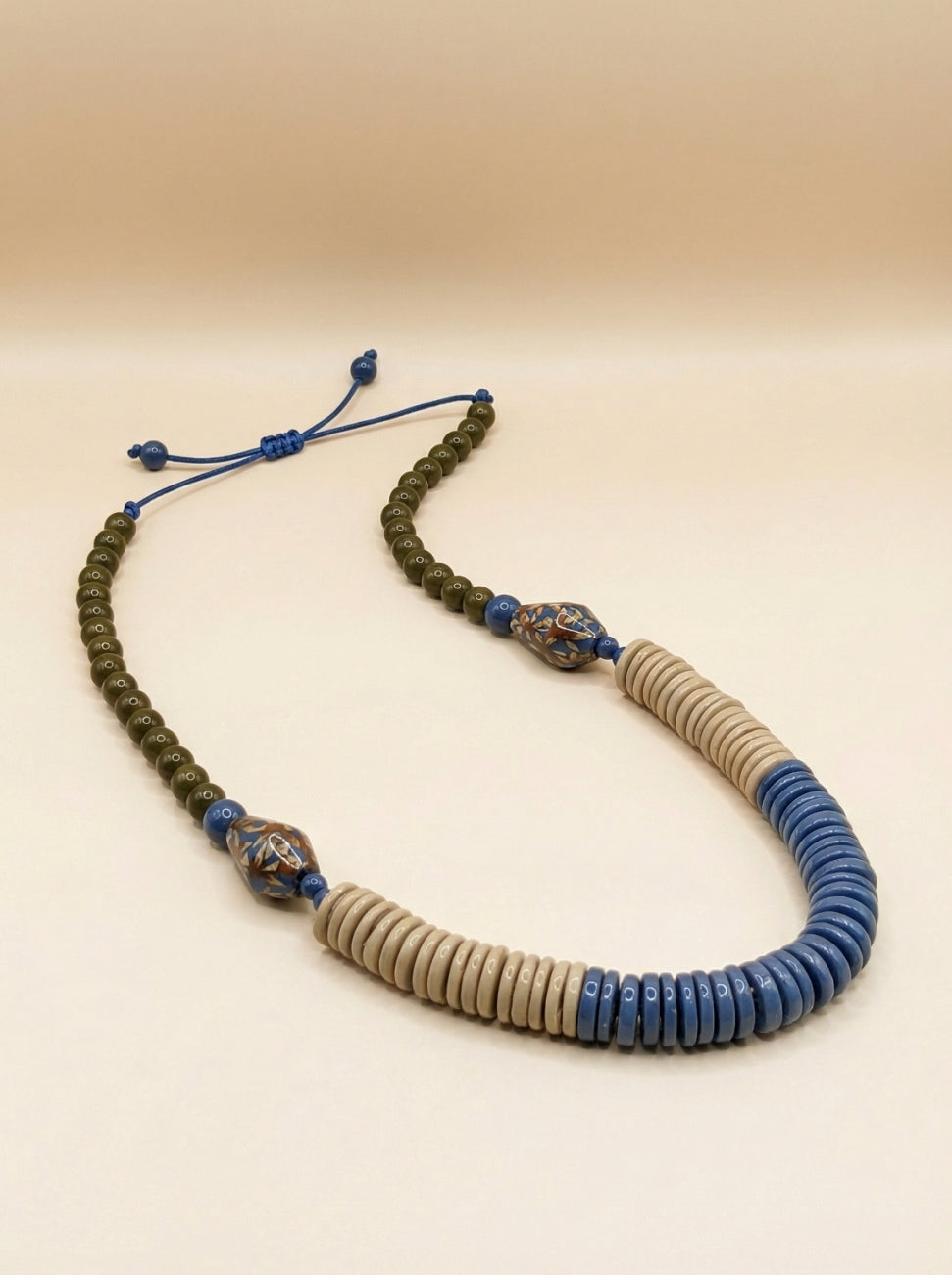 A beaded necklace featuring a dark olive green beaded section, a central section of light beige round beads, a blue beaded section, and two decorative blue and metallic spacer beads on a beige background.