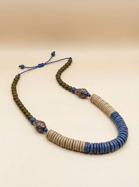 A beaded necklace featuring a dark olive green beaded section, a central section of light beige round beads, a blue beaded section, and two decorative blue and metallic spacer beads on a beige background.