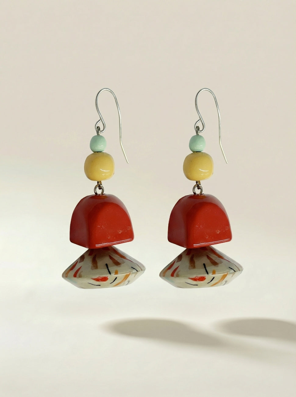 A pair of bohemian dangle earrings featuring a red dome-shaped bead, a marbled white and orange fan-shaped bead, a yellow bead, and a mint green bead on silver French hook wires